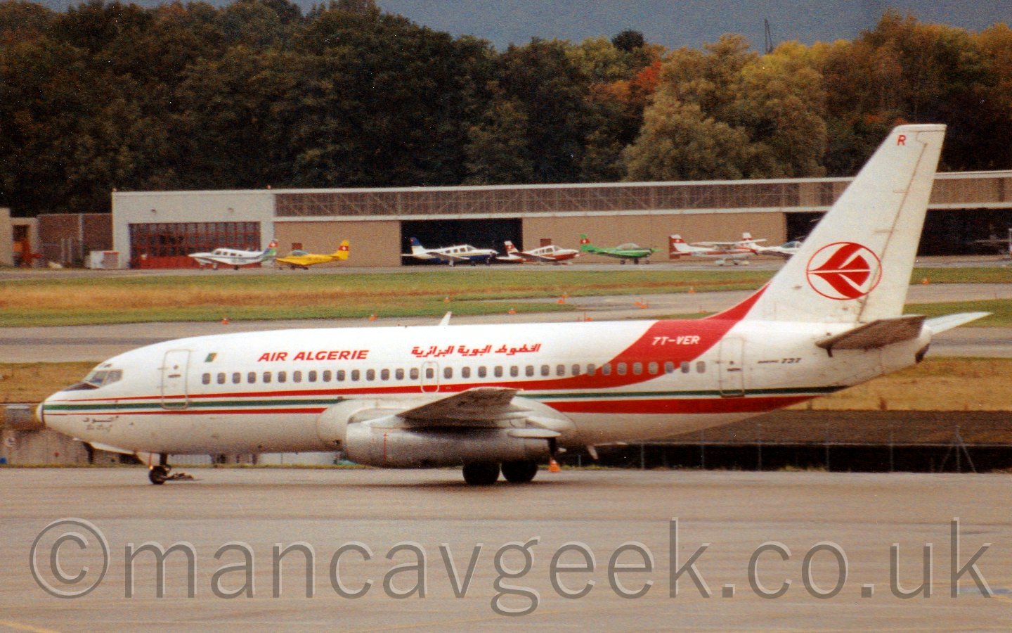 Side view of a twin engined jet airliner taxiing from right to left. The plane is mostly white, with a red/green/red cheatline running along the body, the upper red portion sweeping up over the top of the fuselage just in front of the tail, with the registration "7T-VER" overlaid in white. There are red "Air Algerie" titles on the upper forward fuselage in English, repeated in an Arabic script a little bit further back. The tail has the red outline of a circle, with a stylised image of a red flying bird in the middle., and the red letter "P" right at the top. In the background, grass and runways lead up to a brown hangar in the distance, with an array of variously coloured light aircraft parked in front, with tall trees behind.