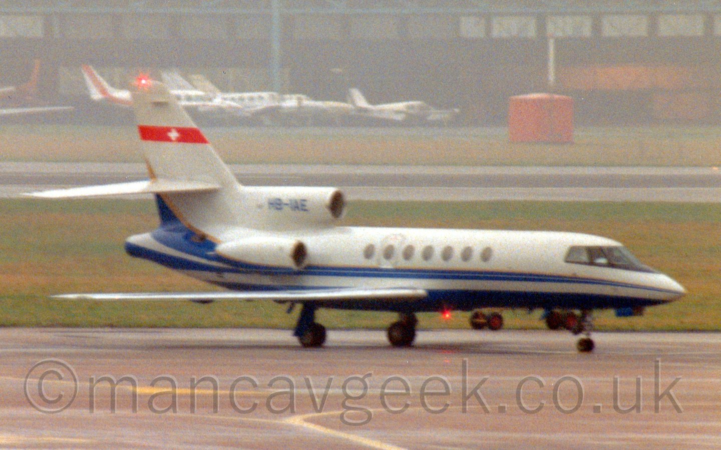 Side view of a 3 engined bizjet parked facing to the right. The plane is mostly white, with a dark blue belly, and a dark blue stripe running along the body, sweeping up arund the lower rear of the engines and tail, with a thin gold stripe on the top edge. There is a red band across the top of the tail whith a white cross in the middle, and the registration "HB-IAE" on the centre engine intake. There is a red beacon shining under the fuselage, with another on the top of the tail. In the background, grass, a runway, and more grass leads up to a large black hangar with a selection of light aircraft parked in front, slowly being swallowed up by mist.