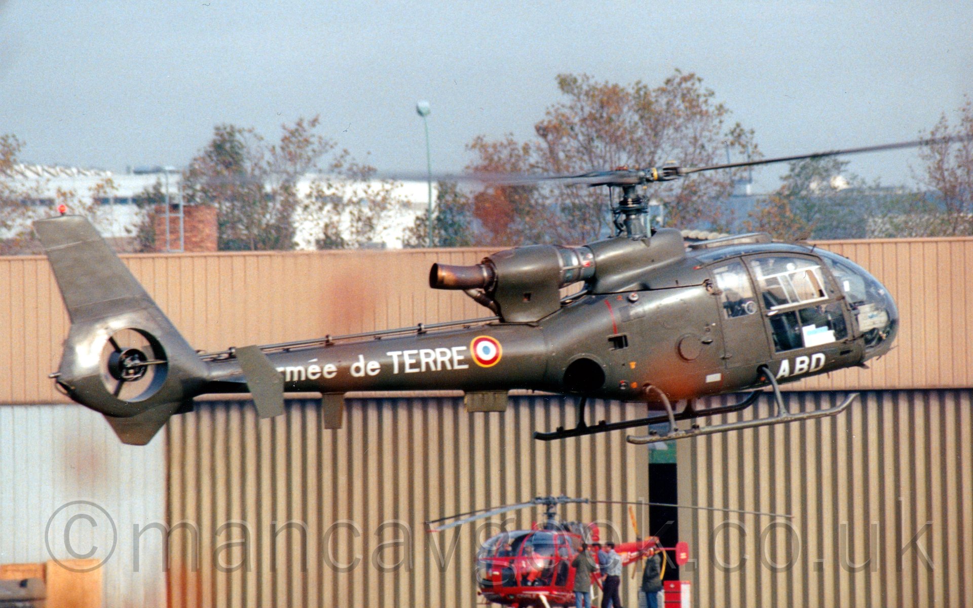 Side view of a military helicopter flying from left to right to land at a heliport. The helicopter is is mostly a drab brown, with a heavily glazed bulbous nose. and a singel engine mounted on the of the top of the fuselage. A long, thick tailbook holds a shrouded fan at the end instead of a normal tail rotor. The letters "ABD" are painted under the cockpit doors, with "Armee de Terre" on the tail boom, next to a red, white, and light blue roundel, outlined in yellow. In the background, a large, sandy-coloured hangar fills most of the frame, with a red helicopter parked in front right at the bottom, with trees and a large white building in the distance, under a hazy grey sky.