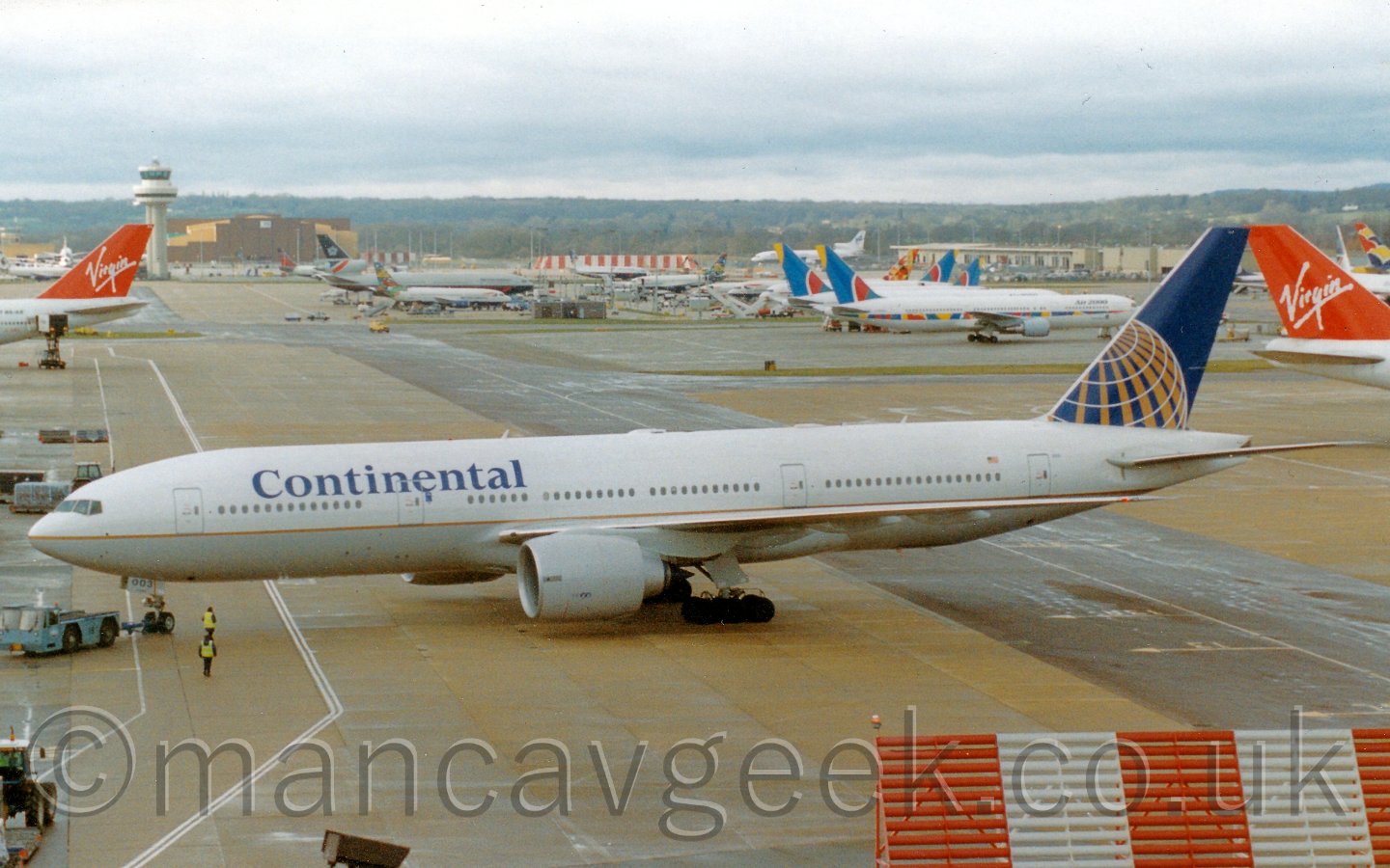 Side view of a white, twin engined jet airliner, being pushed back from it's stand by a blue truck attached to the nose wheel. In the background is a busy scene from an airport, with many planes parked in various areas, under a cloudy white sky.