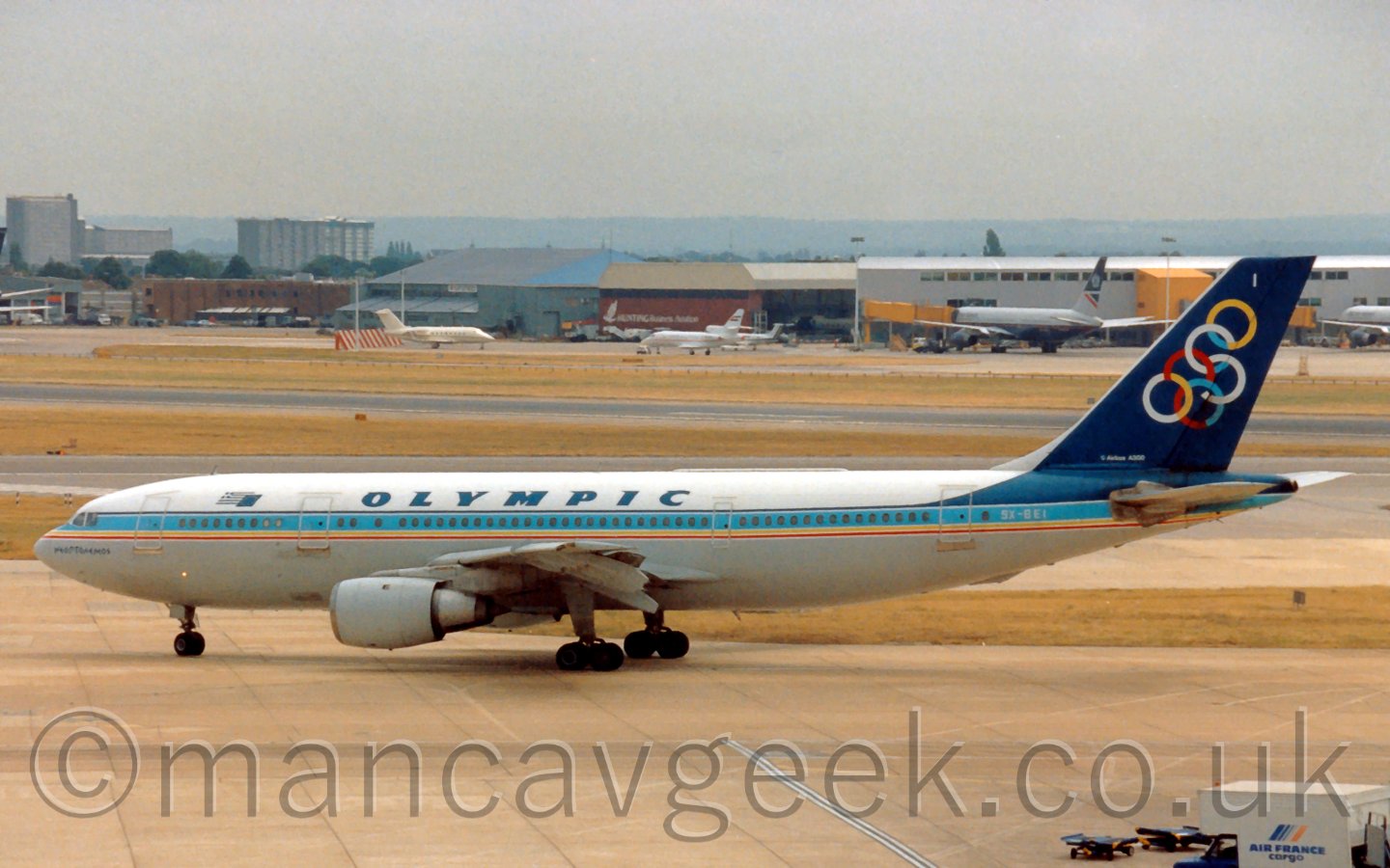 Side view of a twin engined jet airliner taxiing from right to left. The plane has a white top half with a grey belly, seperated with a thick light blue stripe with thinner yellow and red stripes at the bottom, and a dark blue stripe at the top, sweeping up in to the tail. There are blue "Olympic" titles on the upper fuselage. In the background, a grey terminal with yellow airbridges, some attached to grey planes, is on the upper right of the frame, and with a couple of hangars, one blue, one brown, with some bizjets parked in front. Hi-rise buildings in the distance dominate the left of the frame, under a grey sky.