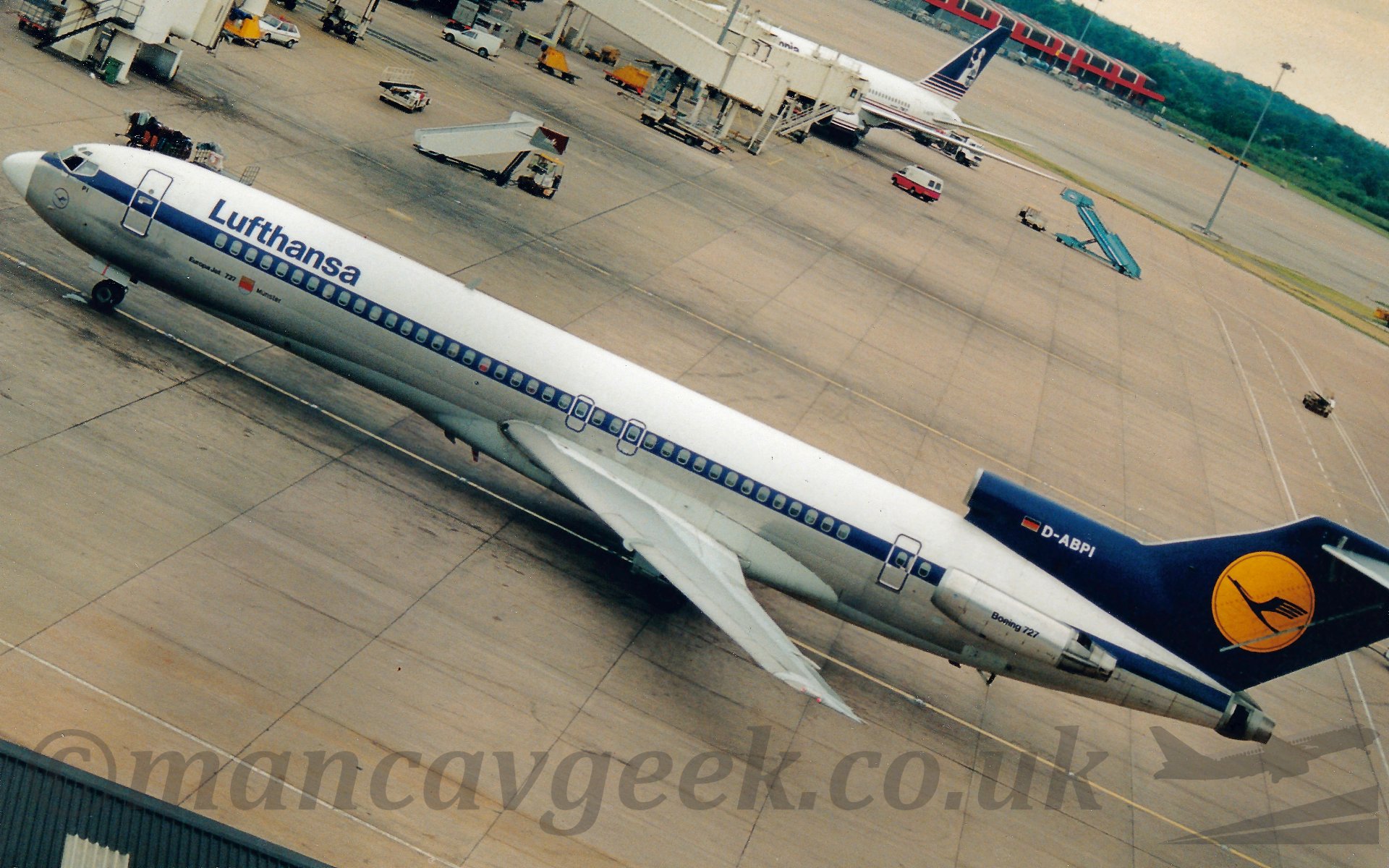 Side view of a 3 engined jet airliner with the engines mounted on the rear fuselage taxiing from right to left, The plane has a white top half and bare metal lower half, seperated by a blue cheatline running over the passenger cabin windows. There are blue "Lufthansa" titles on the upper forward fuselage, and black "Boeing 727" titles on the side mounted engine pods. The tail is a dark blue, with a large yellow circle containing a blue bird with wings outstretched flying from right to left, abnd the registration "D-ADPI" on the centre engine intake. Concrete apron fills most of the rest of the frame, with a couple of white airbridges sticking out, one of which at the top of the frame has a white plane with a blue and white tail attached. In the distance, right at the top of the frame, a red, brown, and white building is at the edge of the apron, with trees behind under a light pink sky.