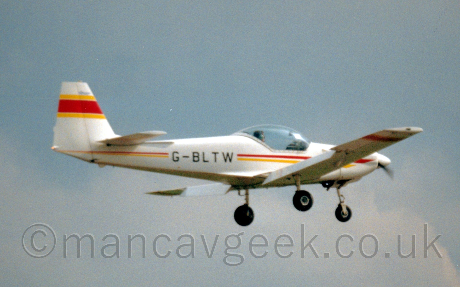 Side view of a single engined light aircraft flying from left to right at a low altitude. The plane is mostly white, with a red and yellow cheatline running along the body, with the registration "G-BLTW" overlaid in black on the rear fuselage. The tail is white, with a yellow-red-yellow stripe running from front to back. The planes cockpit has a large bubble canopy, with the sun reflecting strongly off the front. In the background, grey cloud fills the frame, with hints of blue peeking through.