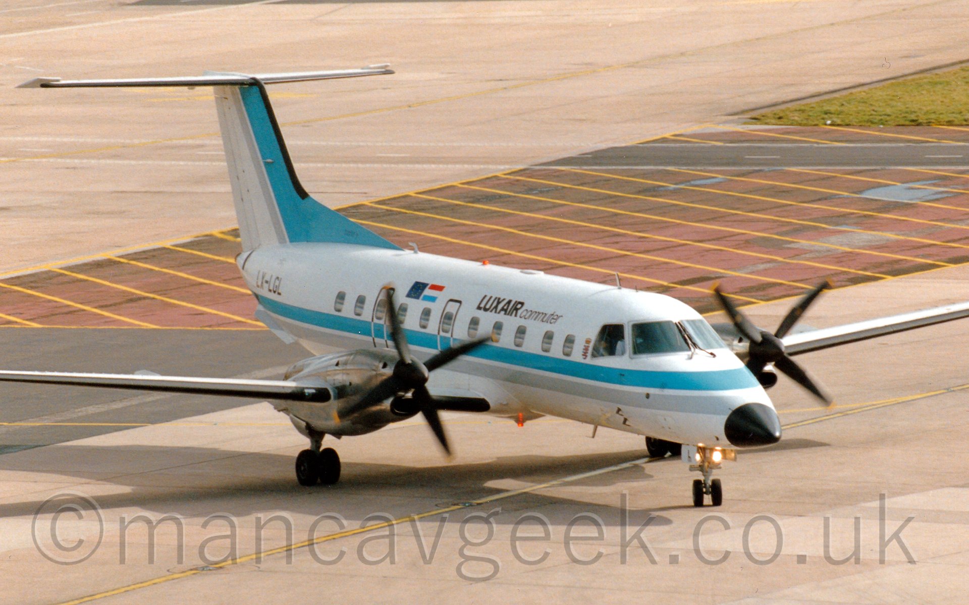 Side view of a twin propellor engined airliner taxiing from left to right, seen from a slightly elevated viewpoint. The plane is mostly white, with a light blue and grey stripe running along the body, repeated diagonally on the tail. There are large "Luxair" and smaller "Commuter" titles in black on the upper forward fuselage, with the flags of both the EU and Luxembourg a bit further back. The registration "LX-LGL" is on the rear fuselage in black. In the background, grey concrete apron fills most of the screen, with red-painted concrete with yellow hatchingdelineating a non-accessible area in the middle.