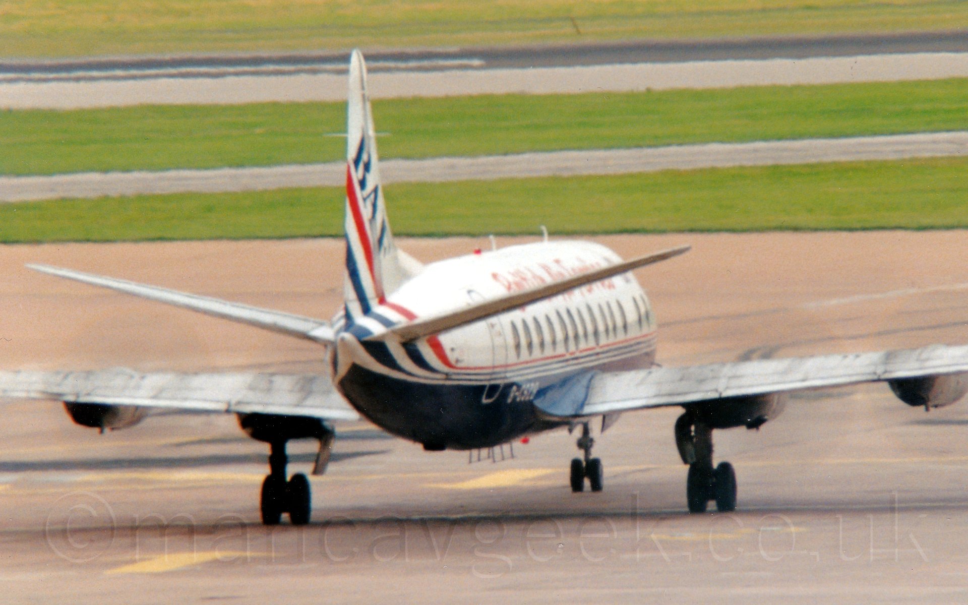 Rear view of a 4 propellor-engined airliner taxiing away from the camera and slightly to the right. The plane is mostly white, with a red and blue stripe separating that from the blue belly. There are red "British Air Ferries" on the upper forward fuselage, and the registration "G-CSZB" on the lower rear fuselage in white. In the background, grey apron leads up to grass, which fills the rest of the frame.