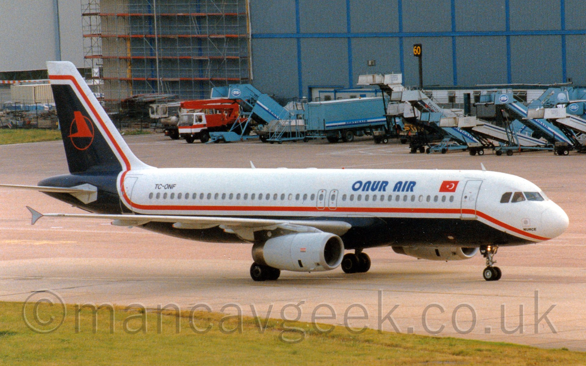 Side view of a twin engined jet airliner taxiing from left to right. The plane is mostly white, with a deep blue belly that sweeps up to fill most of the rear of the tail, and a red cheatlin running along the body from under the nose and up into the tail, seperating the 2 colours. There are pale blue "Onur Air" titles on the upper forward fuselage, just aft of a Turkish flag (red field with a white star in the center and white crescent offset it the right), and the registration "TC-ONF" on the upper rear fuselage in black. The tail has the red outline of a circle, containing a stylised image of a red flying bird. In the background, a large blue hangar dominates the rest of the frame, with various airstairs, trucks, trailers, and cranes parked randomly in front.