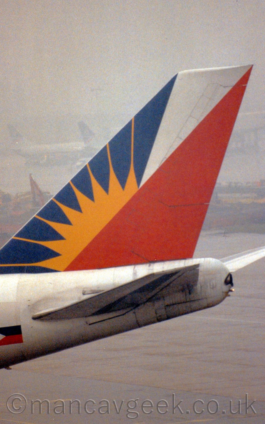 Closeup of the tail of a jet airliner parked facing to the right. The plane is mostly white, with a pair of triangle on the tail, one blue, the other, red, the latter partially covering the former, with an yellow sunburst overlaid on the blue triangle, peeking out from bahind the red. In the background, some construction equipment and a couple of other planes are vaguely discernable in the distance, having been all but swallowed up by fog.