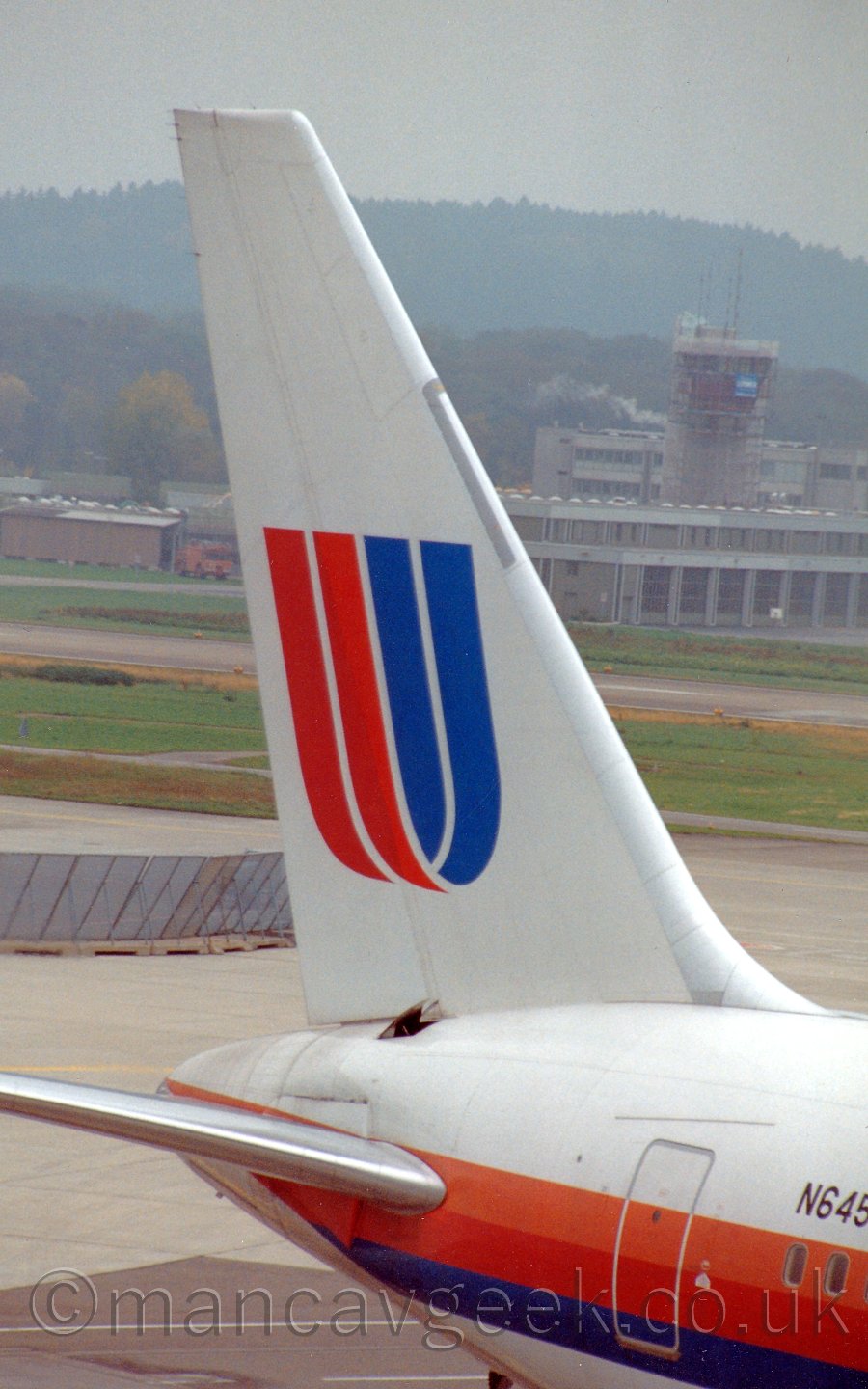 Closeup of the tail of a jet airliner parked facing just off to the right of the camera. the plane is mostly white, with an orange, red, and blue cheatline running along the body, and the registration "N645UA" on the upper forward fuselage in black, the last 2 letters cut off by the side of the frame. The tail is white, with a large letter "U" which looks too be shaped from a piece of ribbon, red and orange on one side, blue on the other. There is a small hatch open on the upper rear fuselage, in the middle of the join with the tail. In the background, taxiways and a runway are seperated by grass, with a large grey building on the far side, and trees on a hill beyond that, meeting hazy grey clouds.