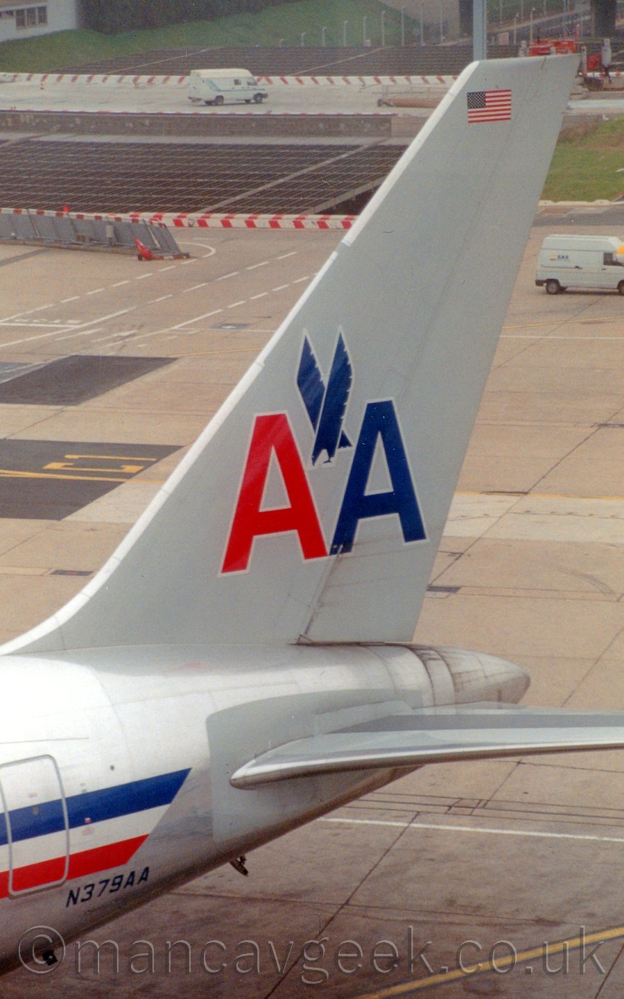 Closeup of the tail of a jet airliner parked facing to the right and angled slightly towards the camera. The plane is mostly grey, with a silver belly, and a blue, white, and red stripe running along the body. There are a pair of capital letter "A"s on the tail, one red, one blue, with a stylised image of a blue stooping eagle between them. In the background, taxiway fills most of the frame, with a metal mesh covering a road tunnel under the airport towards the top of the frame.