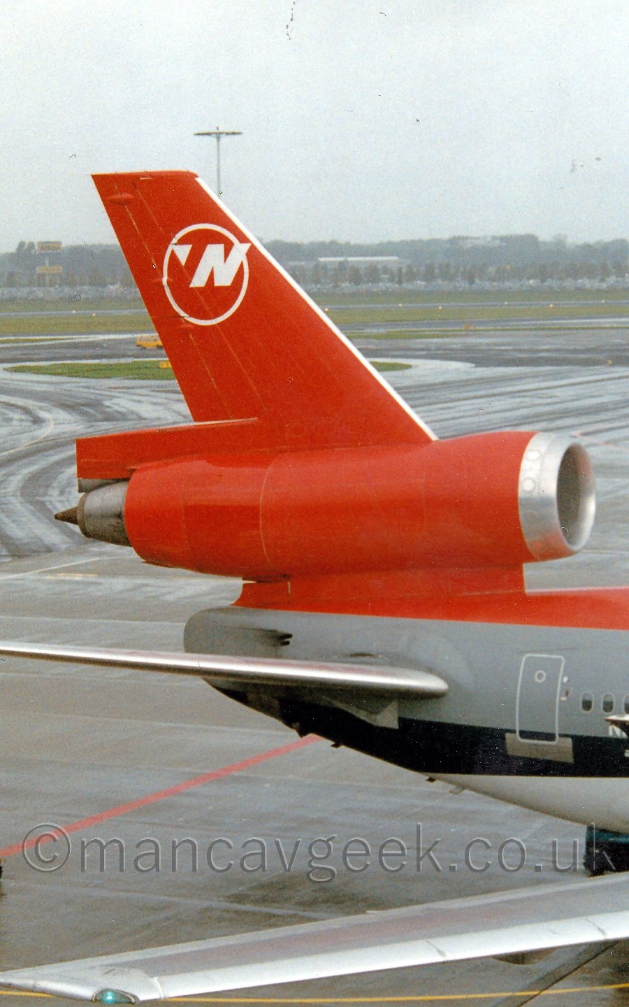 Closeup of the tail of a three engined jet airliner parked facing to the right. The plane has a red top, with a white belly, and a thick grey and black stripe running along the body. The tail is red, with the white outline of a circle, with a white letter "N" overlaid i the top right, and a white triangle pointing to the NorthWest in the top left. In the background, black taxiways fill most of the frame, with grass beyond that leading to trees in the distance, slowly vanishing in to mist.