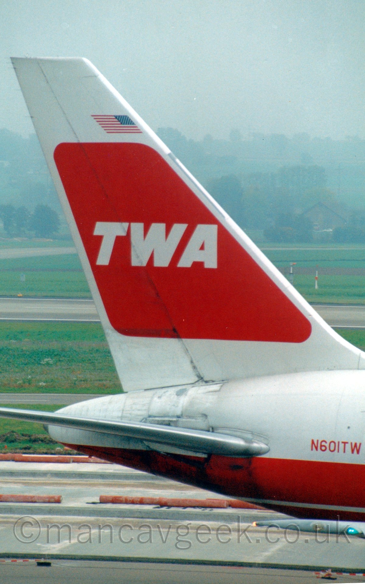 Closeup of the tail of a jet airliner taxiing from left to right. The plane is mostly white, with a pair of red cheatlines running along the middle of the body. The registration "N601TW" is on the upper rear fuselage in red. The tail is white, with red splotch in the middel containing the white letters "TWA", with a small, reversed United States flag just above. In the background, taxiways and runways lead off intop the distancewith trees in the distance slowly vanishing into the hazy grey sky.