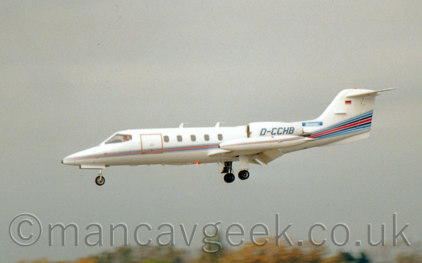 Side view of a white, twin engined bizjet flying from left to right at a low altitude, with undercarriage extednded, flaps deployed from behind the wings, and the nose pointed down slightly, suggesting it is just about to land, with trees at the bottom of the frame, and grey sky filling the rest.