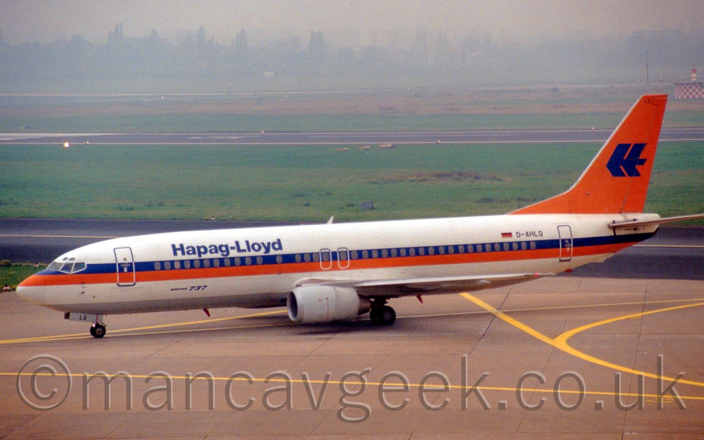 Side view of a white twin jet airliner with a blue and orange stripe running along the body with blue "Hapag-Lloyd" titles on the upper forward fuselage, and an orange tail with a stylised letter "H", taxiing from right to left, with large areas of grass and taxiways in the background vanishing in to mist.