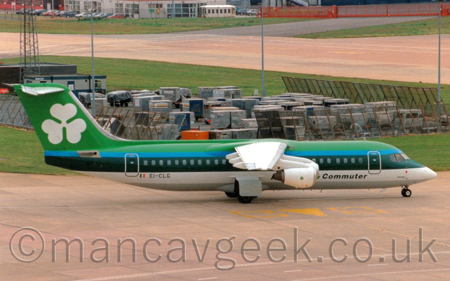 Side view of a green, blue, and white, high-winged, 4 engined jet airliner taxiing from left to right, with lots of luggage containers parked behind metal fences in the background.