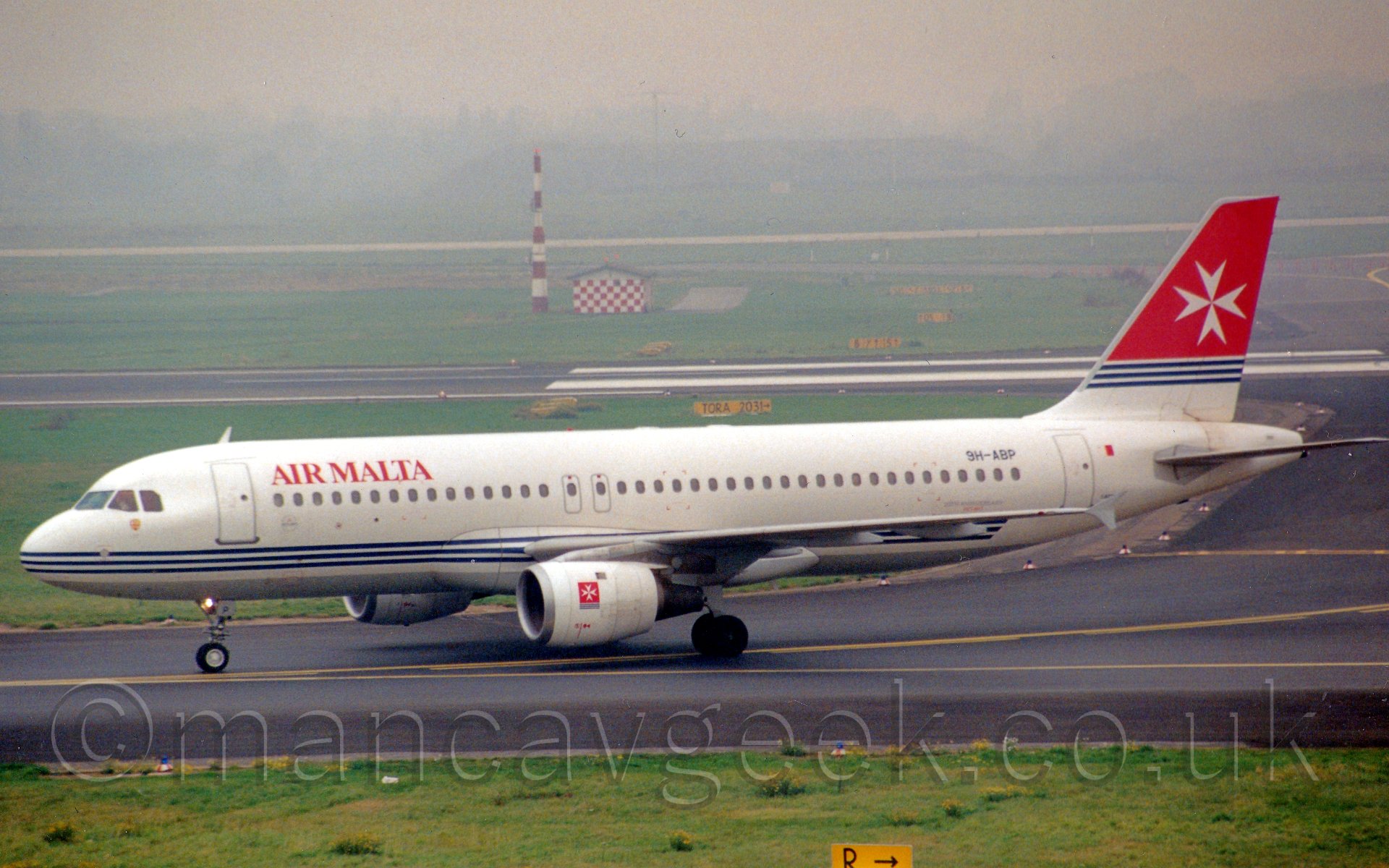 Side view of a twin engined, white jet airliner, taxiing from right to left, with grassed areas and a runway vanishing in to haze in the background.