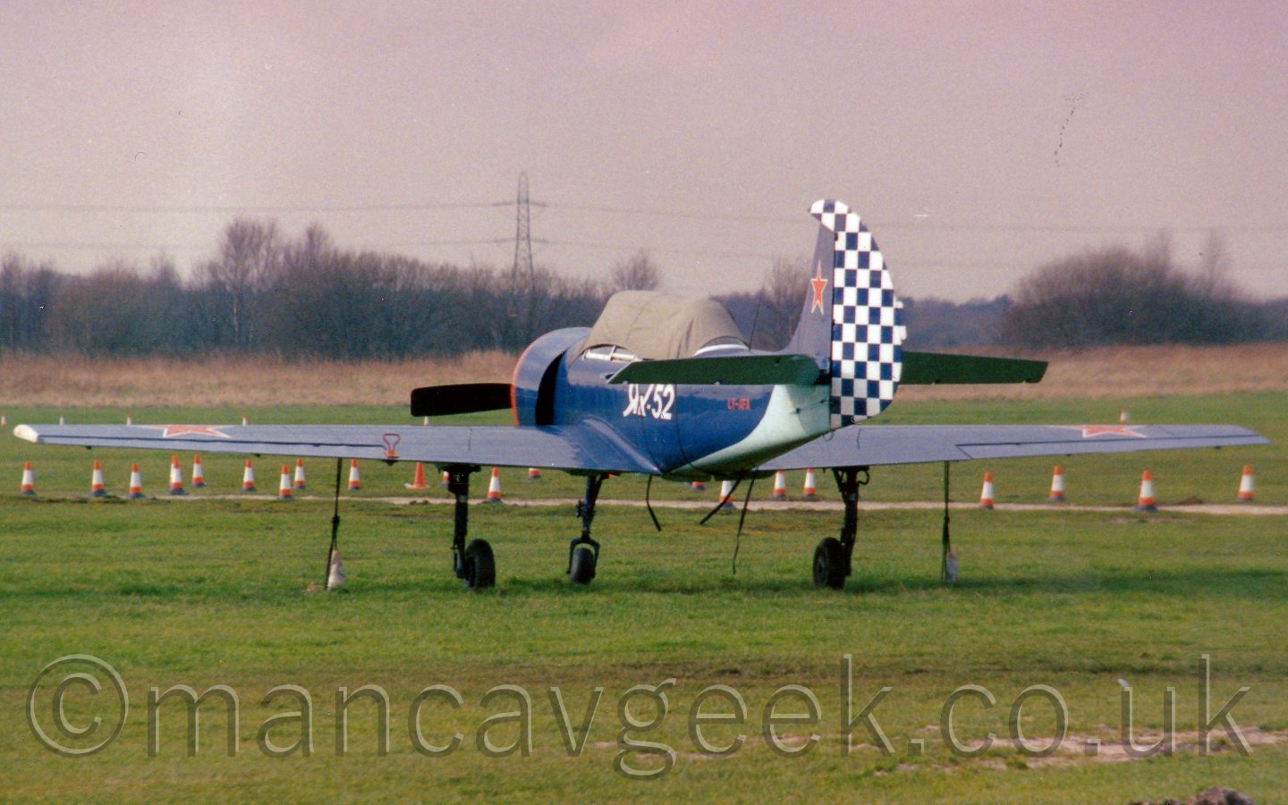 Rear view of a single engined light aircraft with a checkerboard tail parked facing away from the camera on a grass airfield, with trees lining the horizon under a grey sky.