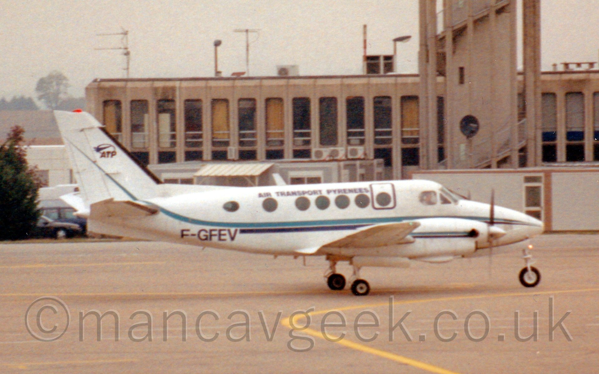 Side view of a white, twin engined bizprop, taxiing from left to right, with a sandy-brown terminal building in the background.