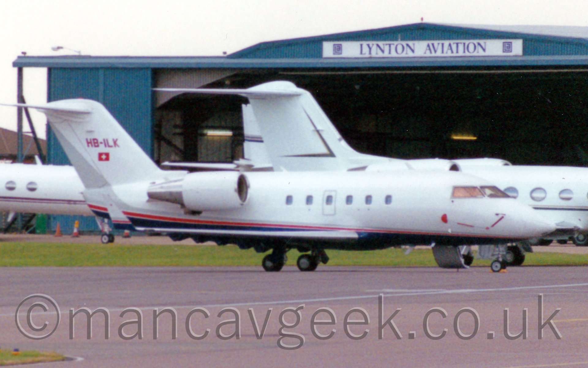 Side view of a white and blue twin engined bizjet parked facing to the right, in front of a large blue hangar with it's doors wide open, surrounded by several other white bizjets.
