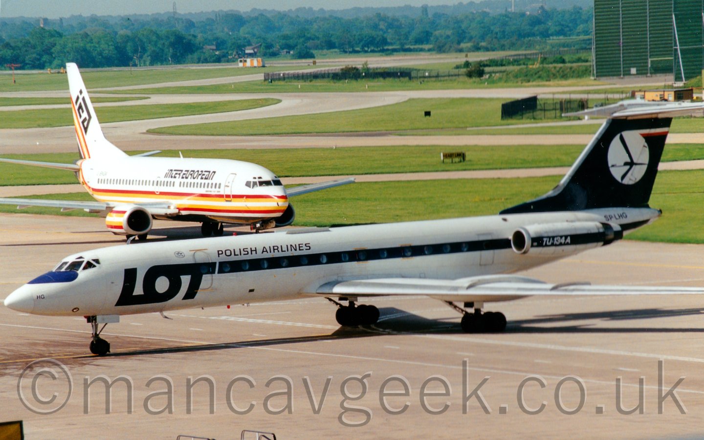 Side view of a white and blue twin engined jet airliner, with the engines mounted on the rear fuselage, taxiing from right to left, with another airliner taxiing towards it in the background, and grass and taxiways leading off to trees in the far distance.
