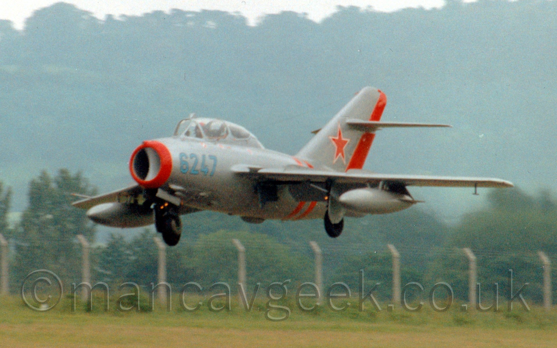 Side view of a silver and red, single engined jet fighter, flying from left to right at a very low altitude, with undercarriage extended and flaps deployed from the rear of the wings, suggesting it is coming in to land, with trees and bushes vanishing into hazy grey sky in the background.