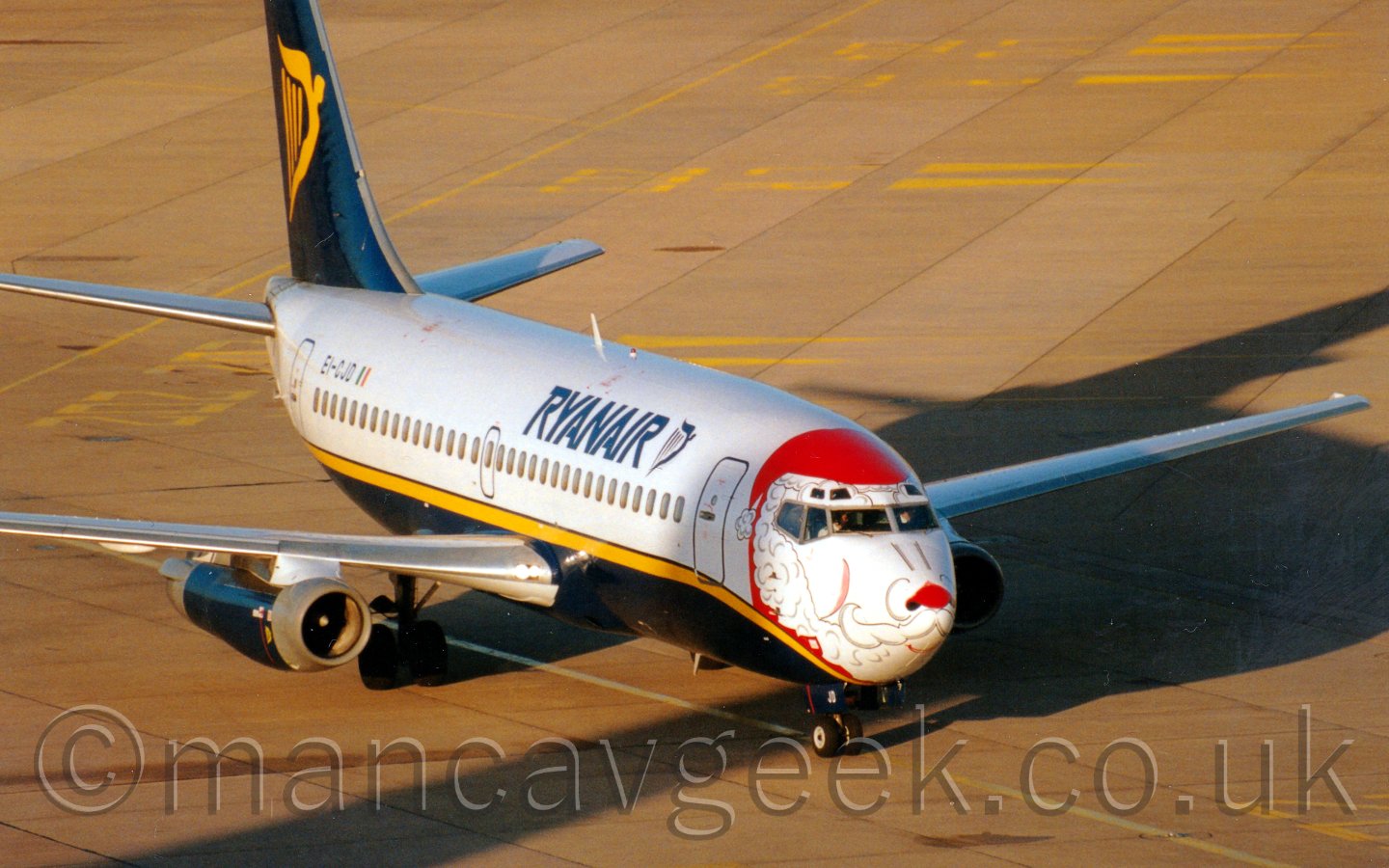 Side view of a white and blue, twin engined jet airliner, with blue "Ryanair" titles on the forward fuselage, and the face of a white bear wearing a red Santa hat and red nose painted around the cockpit, taxiing from left to right, in bright sunshine.