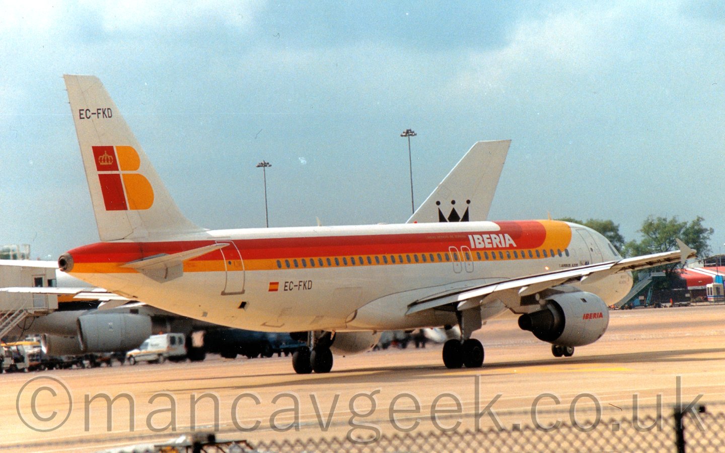 Side view of a white, orange, orange, and yellow, twin engined jet airliner, taxiing from left to right and slightly away from the camera, with another white plane behind it, under a blue sky with bits of white fluff.