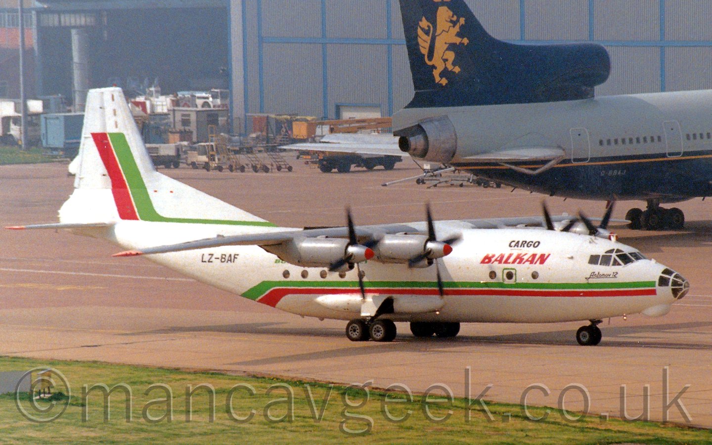 Side view of a white, green and red high-winged, 4 propellor-engined cargo plane taxiing from left to right, with a larger grey and blue plane parked by a grey hangar in the background.