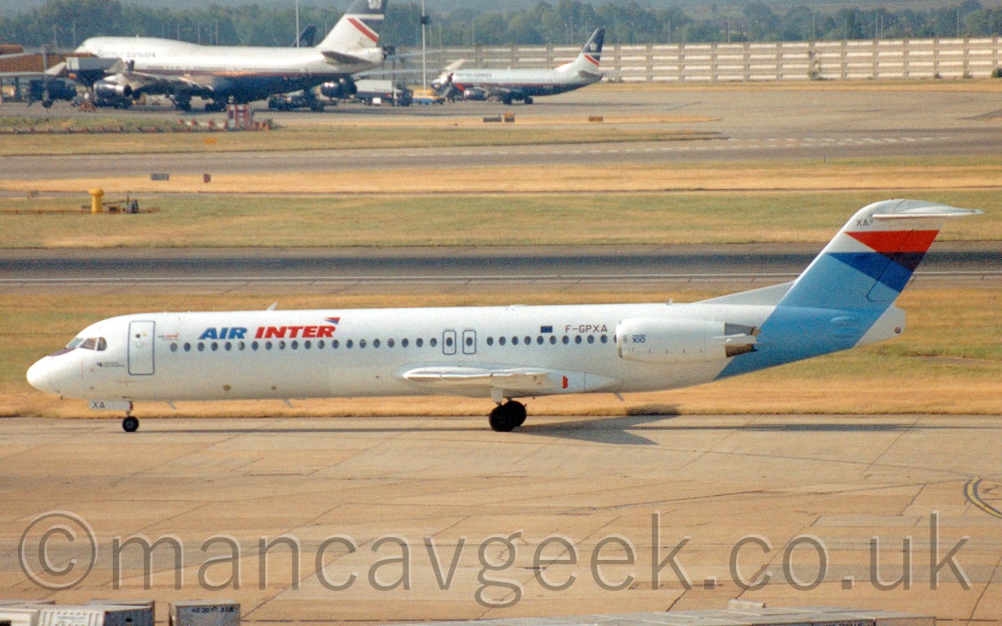 Side view of a white, blue, and red, twin engined jet airliner, with the engines mounted on the rear fuselage, taxiing from right to left, with a coupple of dark blue and grey on the far side of a runway.