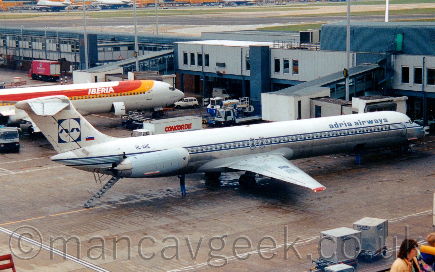 Side view of a white and blue, twin engined jet airliner with the engines mounted on the rear fuselage, parked facing to the right, with a set of buildt-in airstairs deployed from the rear, between the engines, in front of a terminal building, with a white and orange jet airliner parked next to it.