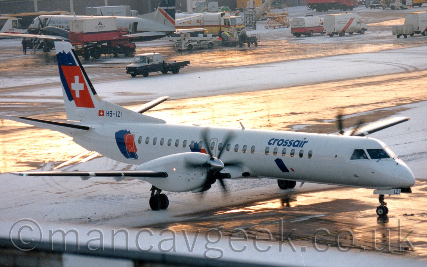 Side view of a white, twin propellor-engined airliner taxiing from left to right, with snow on the ground, and golden light reflecting off some areas that had been cleared on taxiways.