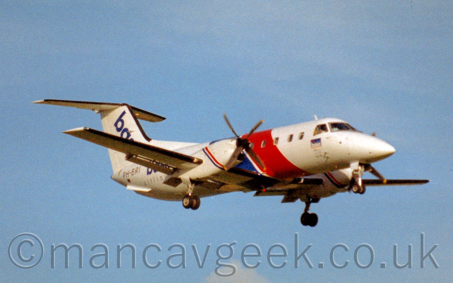 Side view of a white, twin propellor engined alirliner flying from left to right at a low altitude, with wheels extended and flaps deployed from the rear of the wings, obviously about to land at an airport.