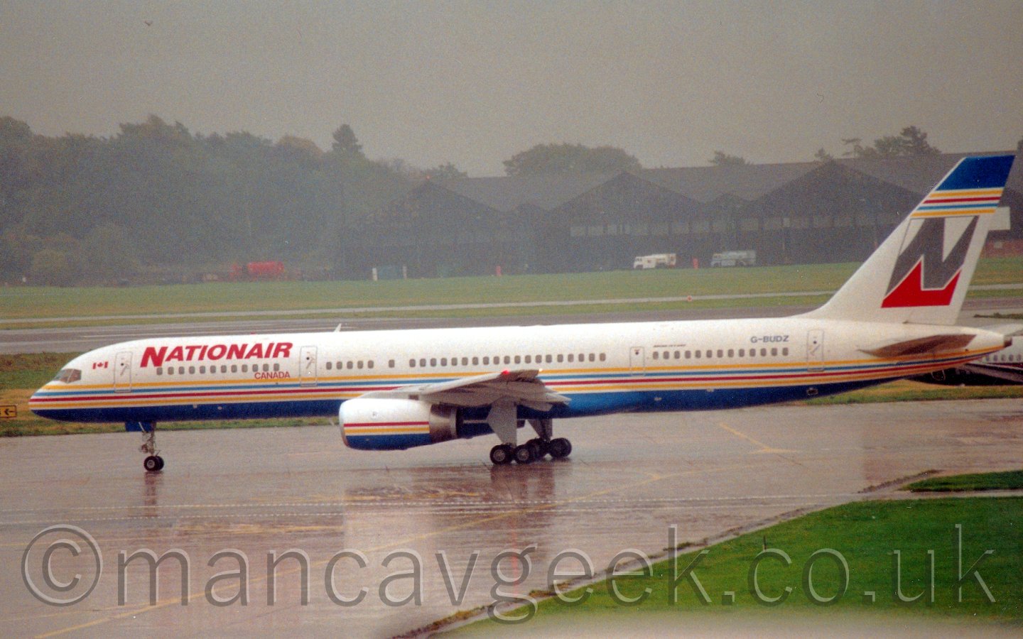 Side view of a white and blue twin engined jet airliner with a white, grey, and red tail, taxiing from right to left at a rather damp airport, under a misty grey sky.