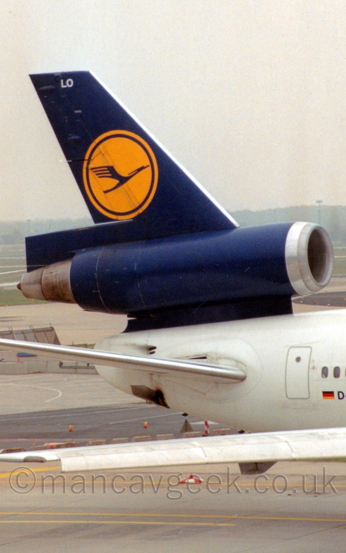 Close up of the blue tail and centre engine of a white 3 engined jet airliner taxiing from left to right, under a hazy white sky.