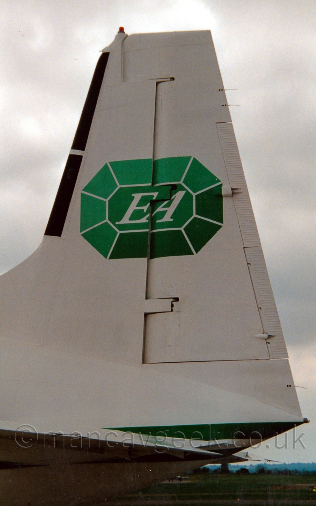Closeup of the tail of a white, twin propellor engined airliner parked facing to the left, with an image of a green precious stone with the letters "EA" in the middle of the tail., under a grey sky.