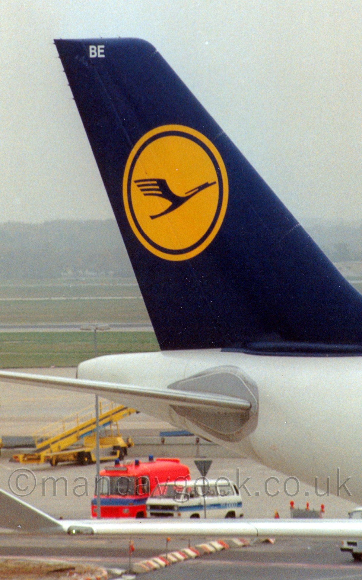 Close up of the white rear fuselage and blue tail of a jet airliner taxiing from left top right, under a misty grey sky.