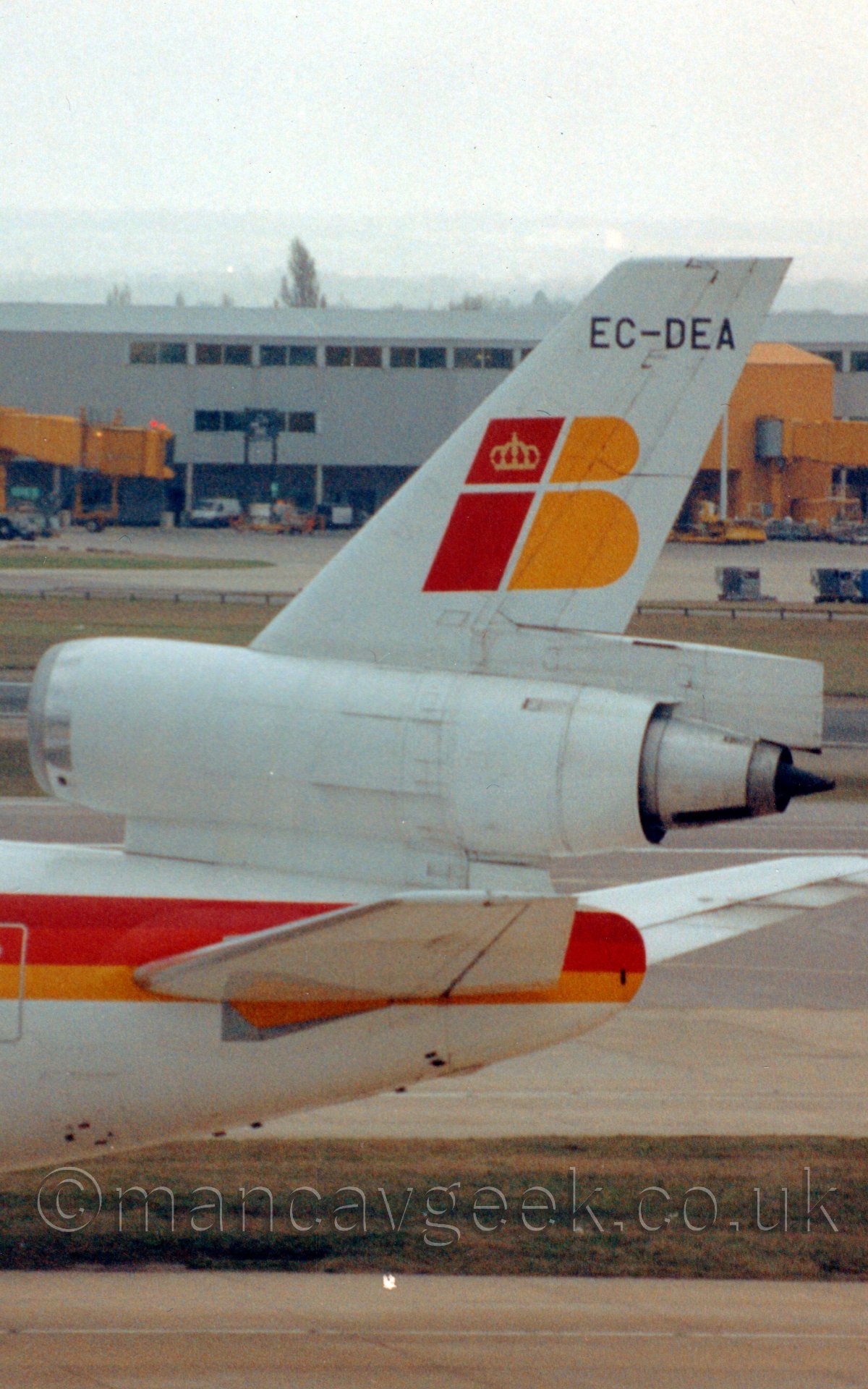 Close up of the white, yellow, and orange rear fuselage and tail of a 3 engined jet airliner taxiing from right to left, under a bright but hazy grey sky.
