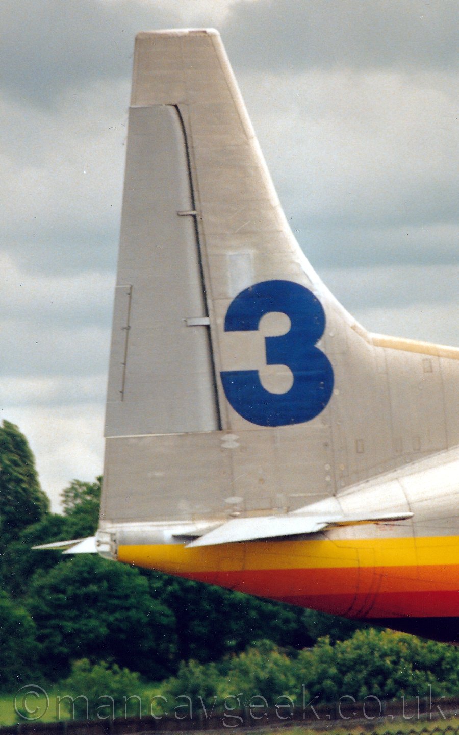 Closup of the silver tail of a silver plane with an yellow, orange, red, and dark blue stripe running along the body, faciong to the right, with trees in the background, under a cloudy grey sky.