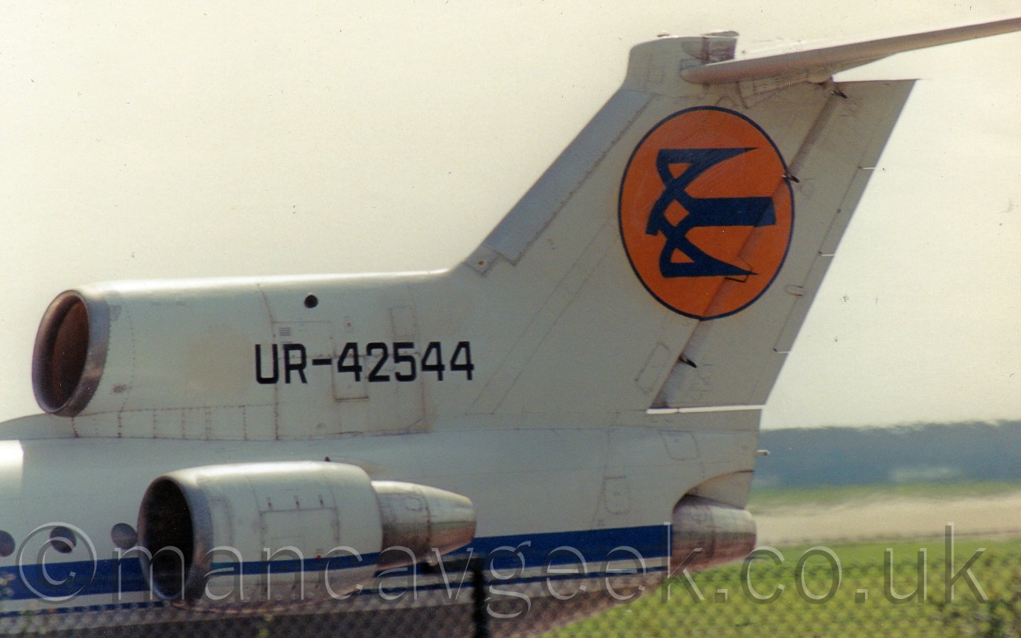 Close up of the tail of a white, 3 engined jet airliner taxing from right to left, under a bright but hazy sky.