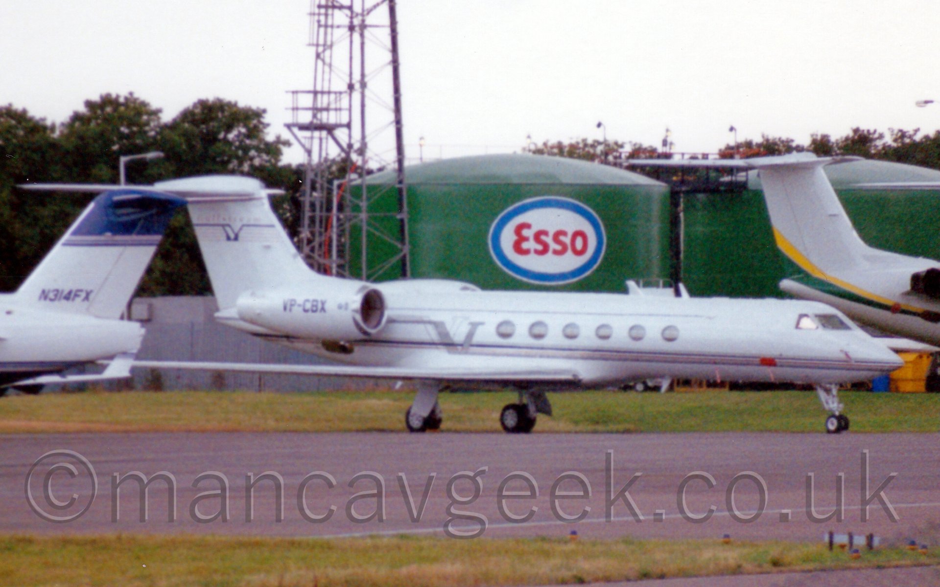 Slightly grainy side view of a white, twin engined bizjet parked facing to the right, with green fuel tanks and the tails of another couple of bizjets in the background.