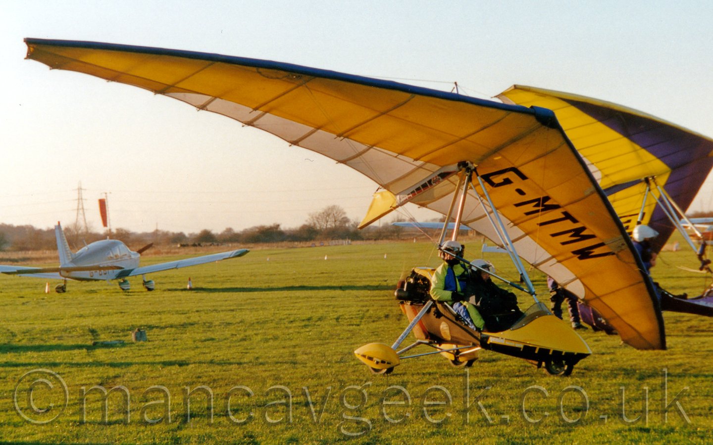 Side view of a yellow microlight, basically a shaped bucket on wheels suspended beneath a large, triangular, fabric-covered wing, with 2 people sitting in tandem in the bicket, and a small engine and propellor mounted on the rear, facing to the righton a grass airfield, under a bright but hazy sky.