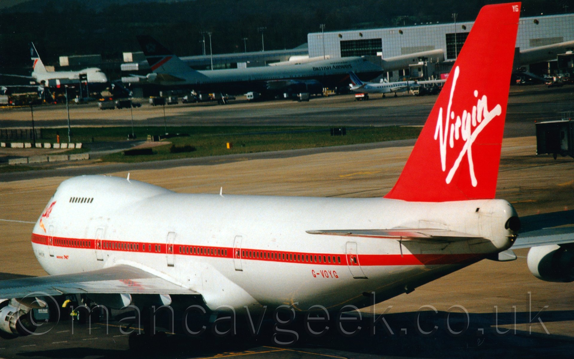 Side view of a white and red, 4 engined, very large jet airliner, taxiing from right to left at a busy airport.