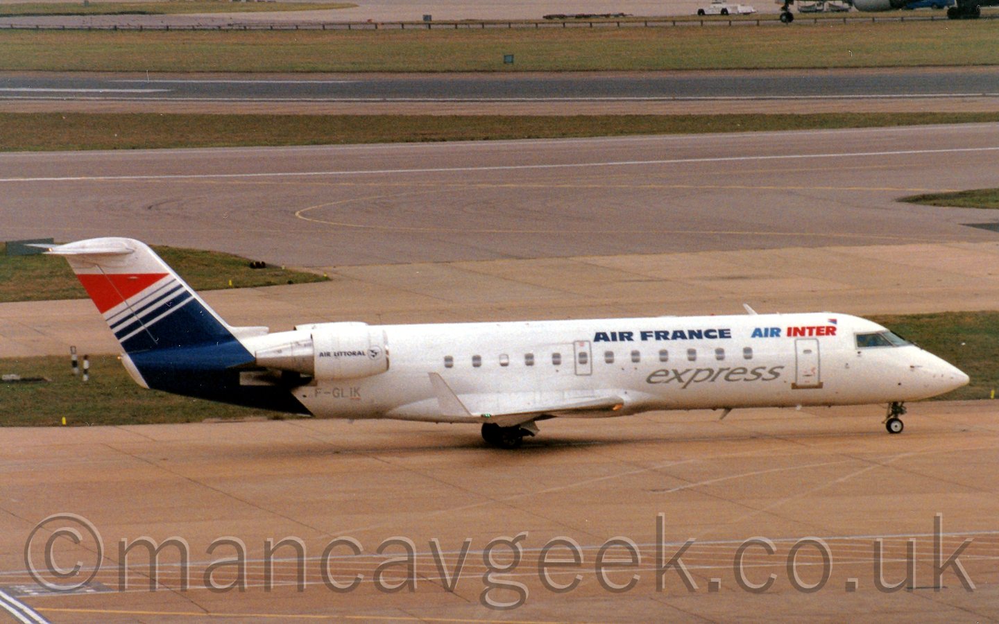 Side view of a white, twin angined jet airliner, with a blue and red tail, and the engines mounted on the rear fuselage, taxiing from left to right.