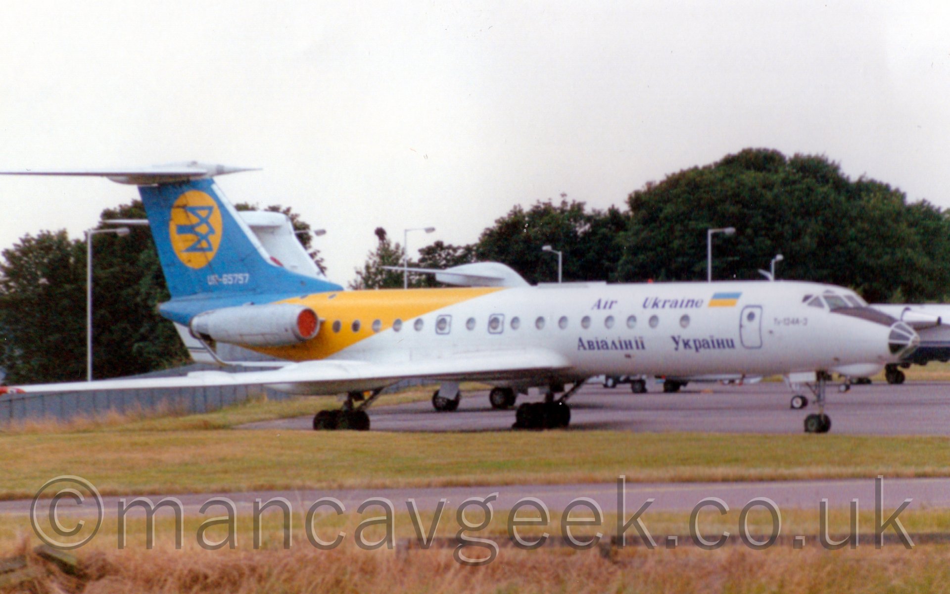 Side view of a white, yellow, and blue, twin engined jet airliner with the engines mounted on the rear fuselage, on the bizjet apron at Luton Airport, some time in the late 1990's.