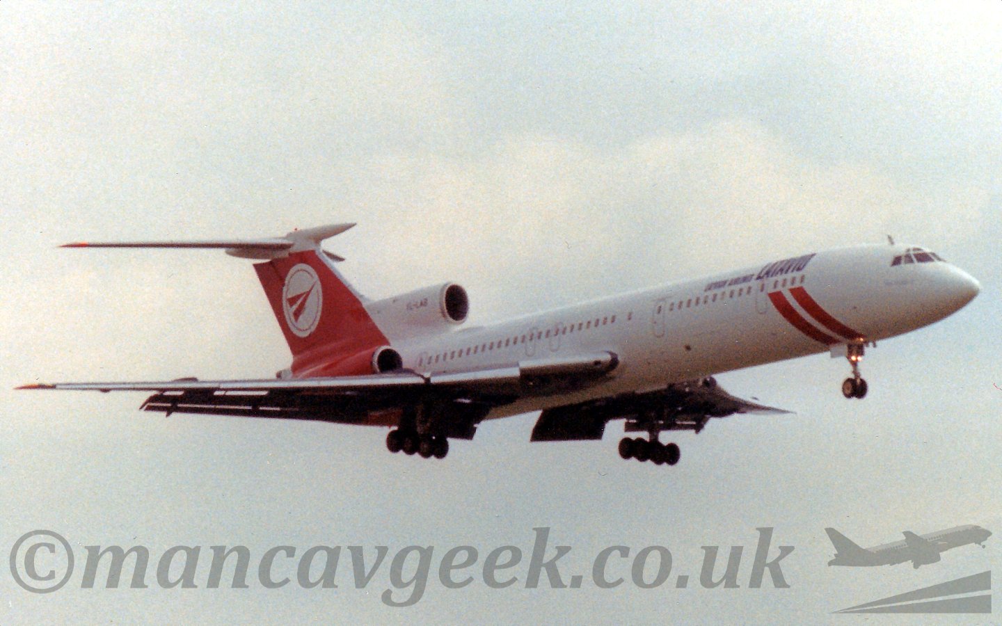 Side view of a white and red, 3 engined jet airliner flying from left to right at a low altitude, with undercarriage lowered and flaps deployed from the rear of the wings, suggesting it is just about to land.