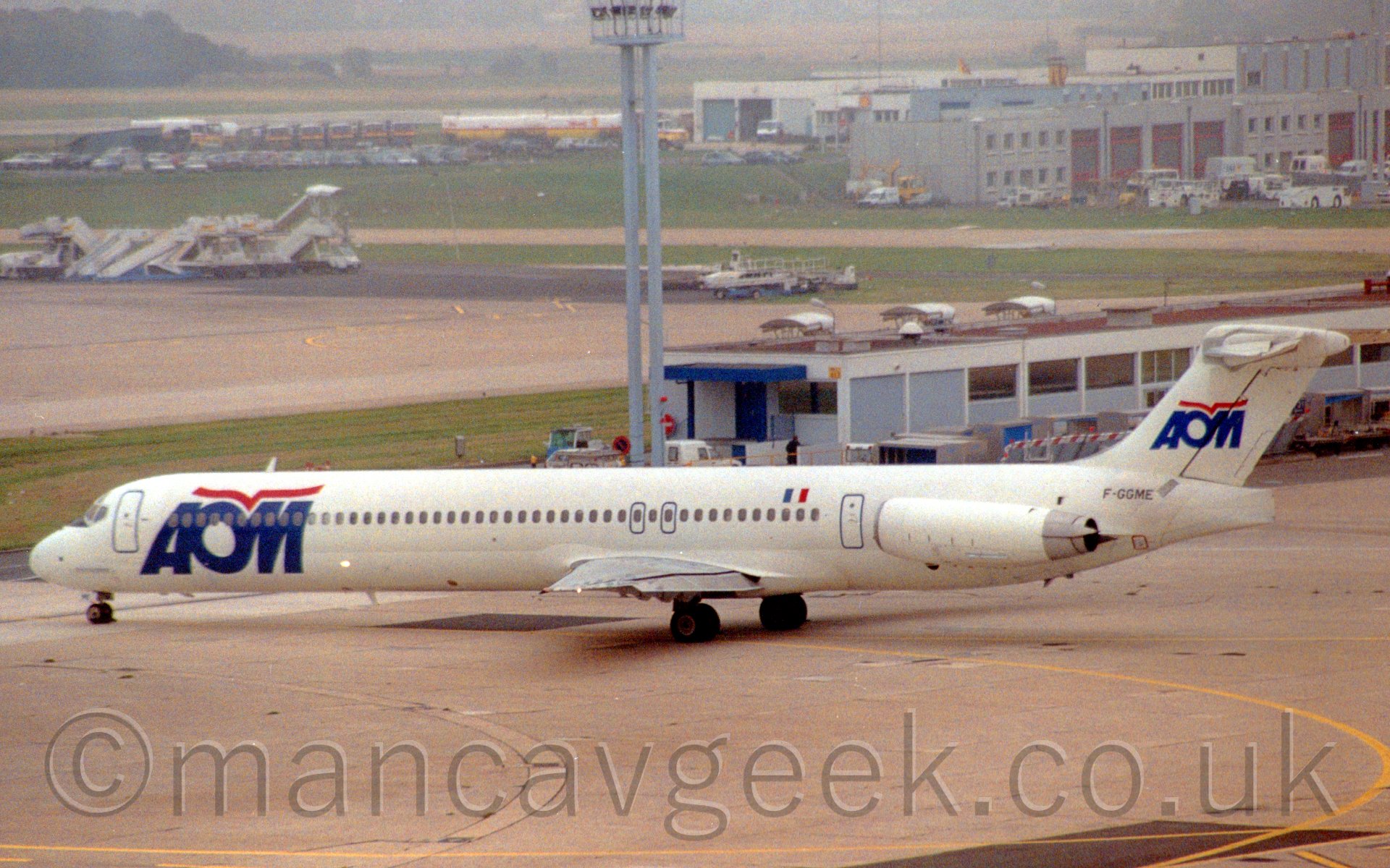Side view of a white, twin engined jet airliner with the engines mounted on the rear fuselage, taxiing from right to left, at a seemingly deserted airport.