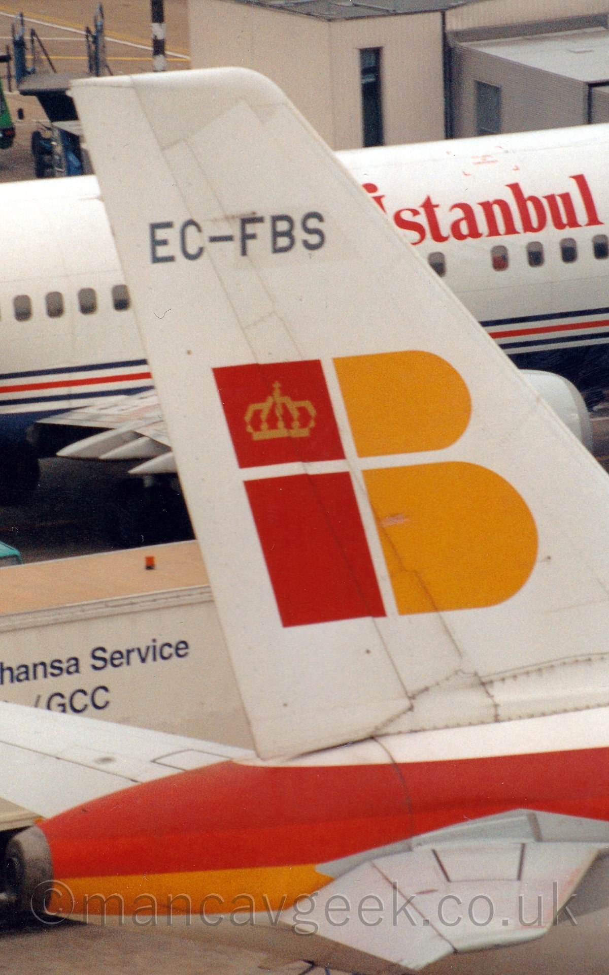 Closeup of the white tail of a jet airliner with the letter "i" and "B" in red and yellow, respectively, in the middle, with another jet airliner in the background.