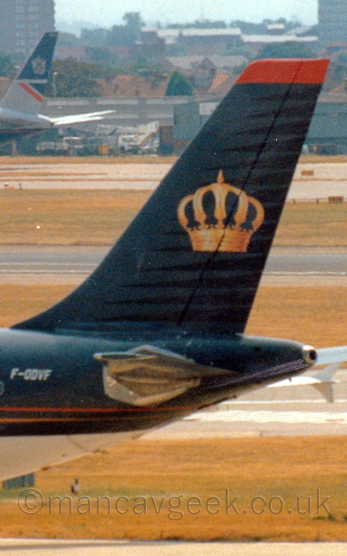 Close up of the tail of a black and grey jet airliner taxiing from right to left, with the tail of another plane in front of an airport terminal building in the background, with housing blocks beyond that.