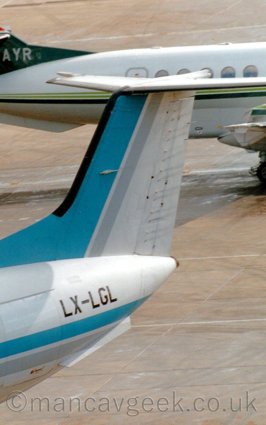 Closeup of the white and blue tail of an airliner taxiing towards the bottom left corner of the frame, with rear fuselage of a white and green plane in the background.