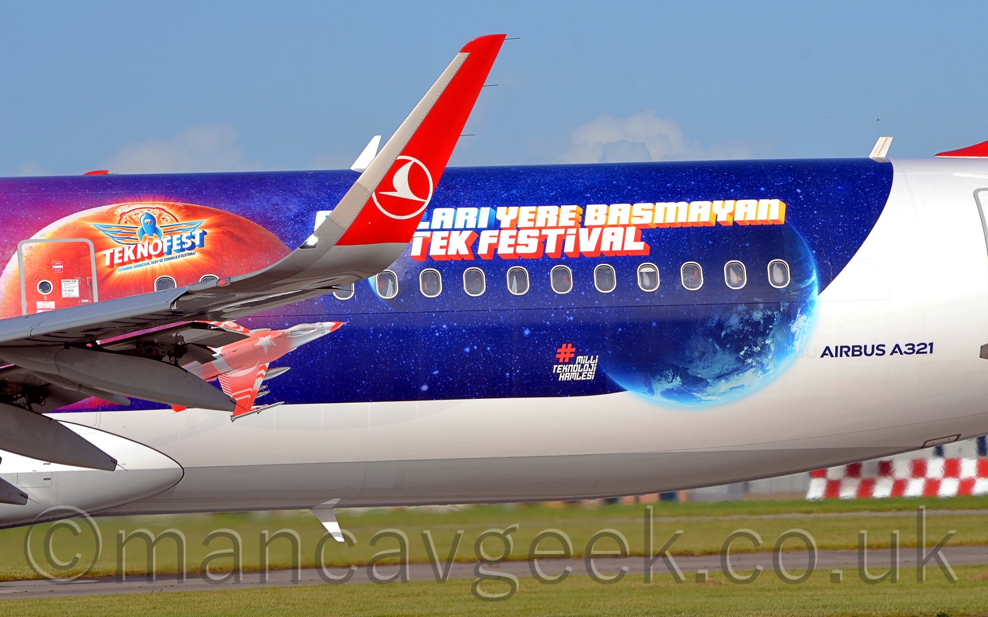 Closeup of the red and white upturnedwingtip of a twin engined jet airliner, with the white and blue rear fuselage behind it advertising a festival in Turkey, under a pale blue sky.