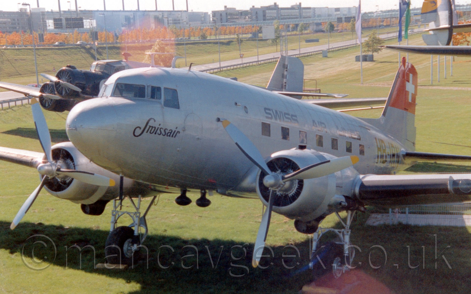 Side view of a bare metal finish, twin propellor-engined World War II-era airliner on display in a grass field with a 3 engined airliner of similar vintage behind it, in bright sunlight.